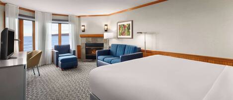 A hotel room with a large bed, a desk with a television, a seating area with a sofa and chairs, a fireplace, and a view of the sea.