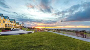 A grassy area with a pathway, benches, and a row of yellow buildings with a sunset in the background.