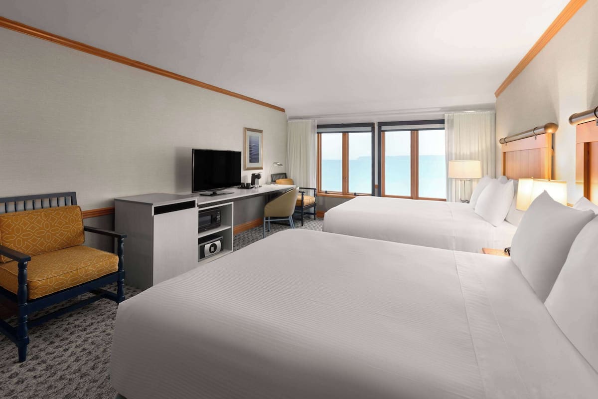 A hotel room with two beds, a desk with a television, a chair, and a view of the sea.