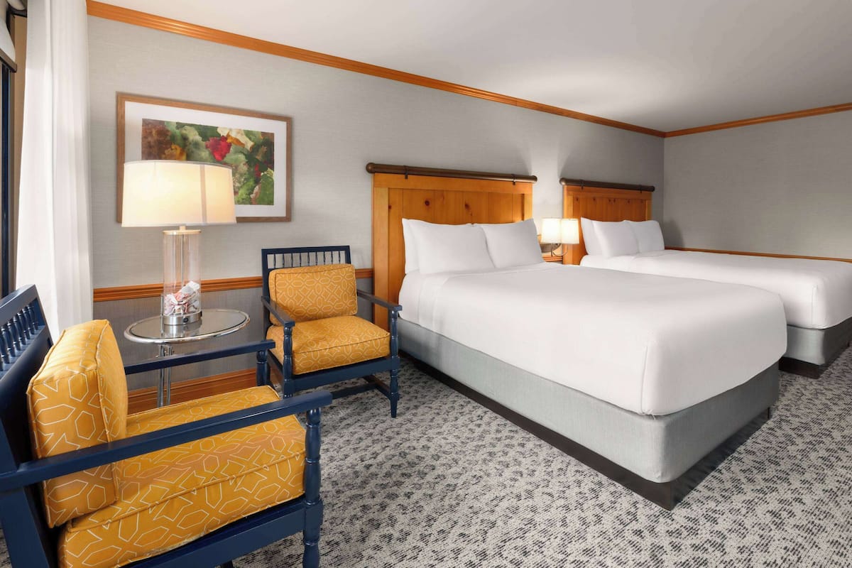 A hotel room with two beds, a chair, a table, and a lamp.