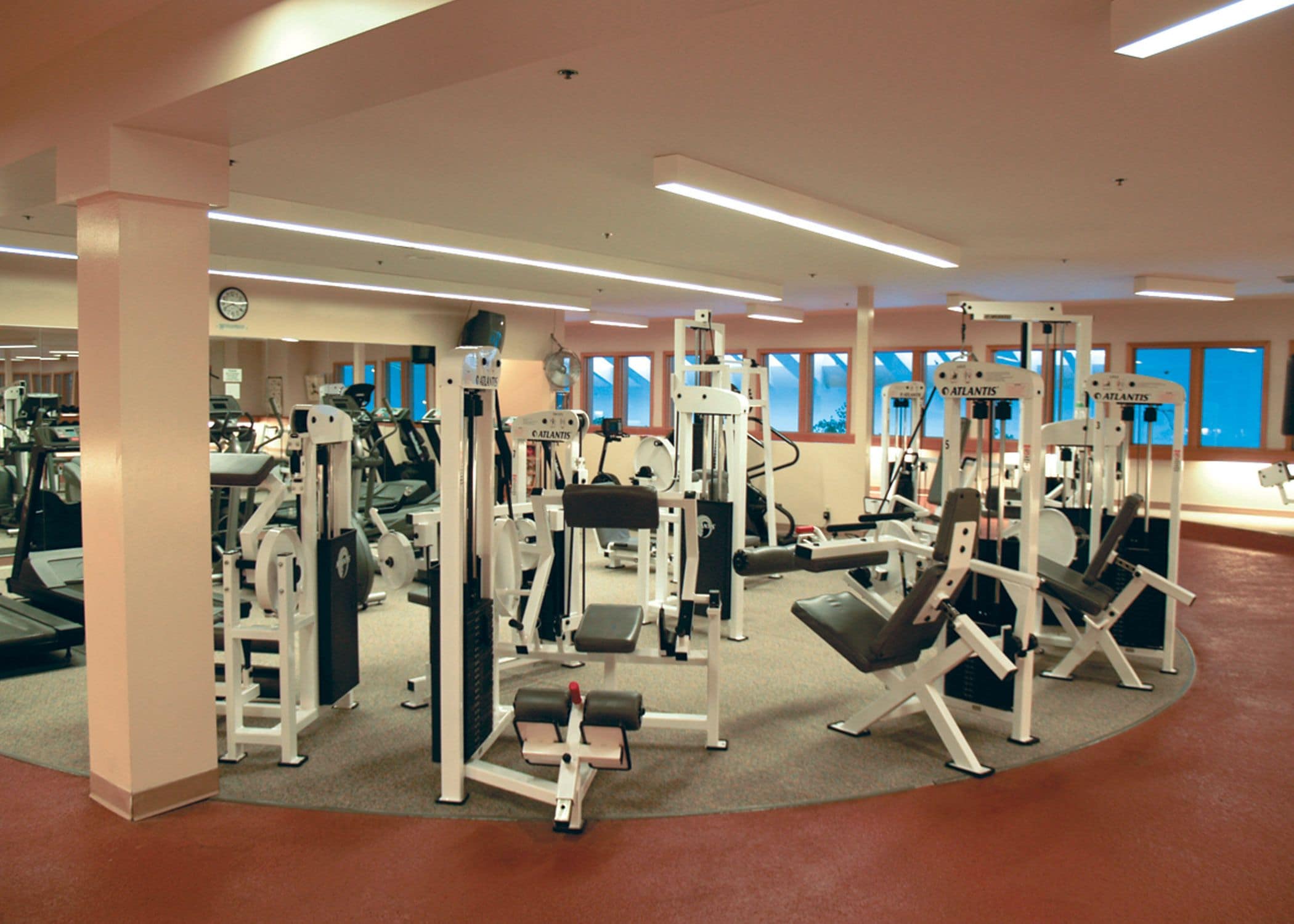 A gym with various exercise machines, large windows, and a clock on the wall.