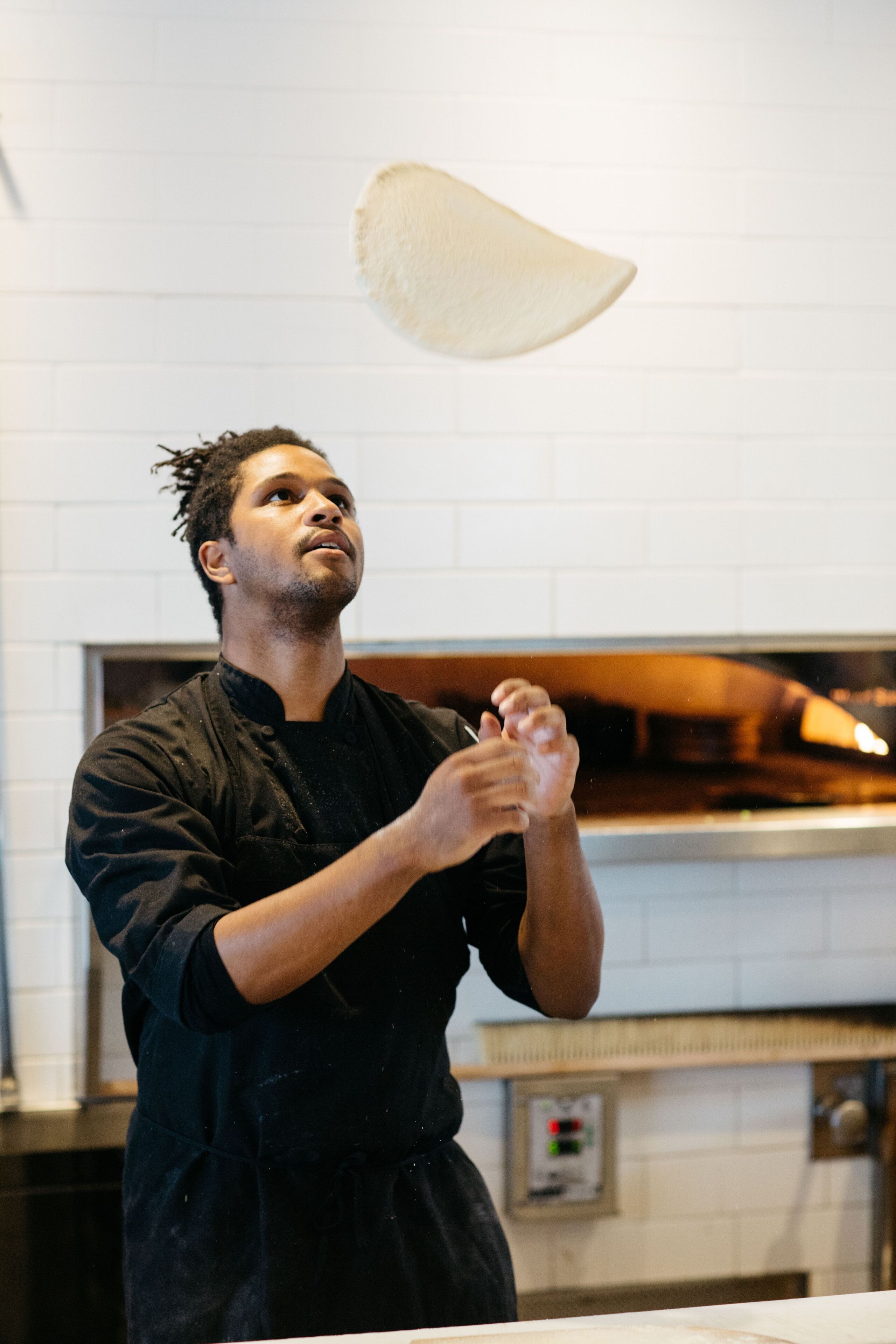 Un chef, vêtu d’un uniforme noir, prépare des plats dans une cuisine aux murs de briques blanches, où l’on aperçoit un four.