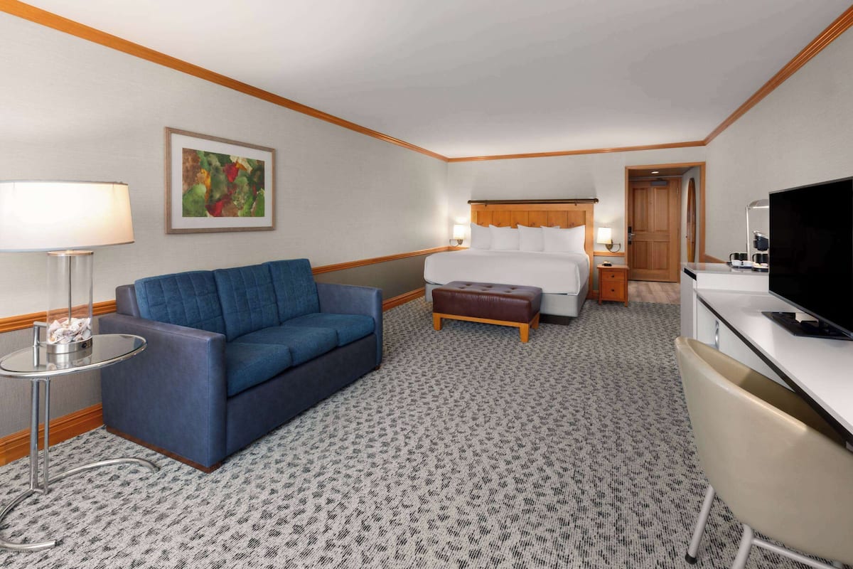 A hotel room with a bed, a blue sofa, a small table with a lamp, a television, and a carpeted floor.