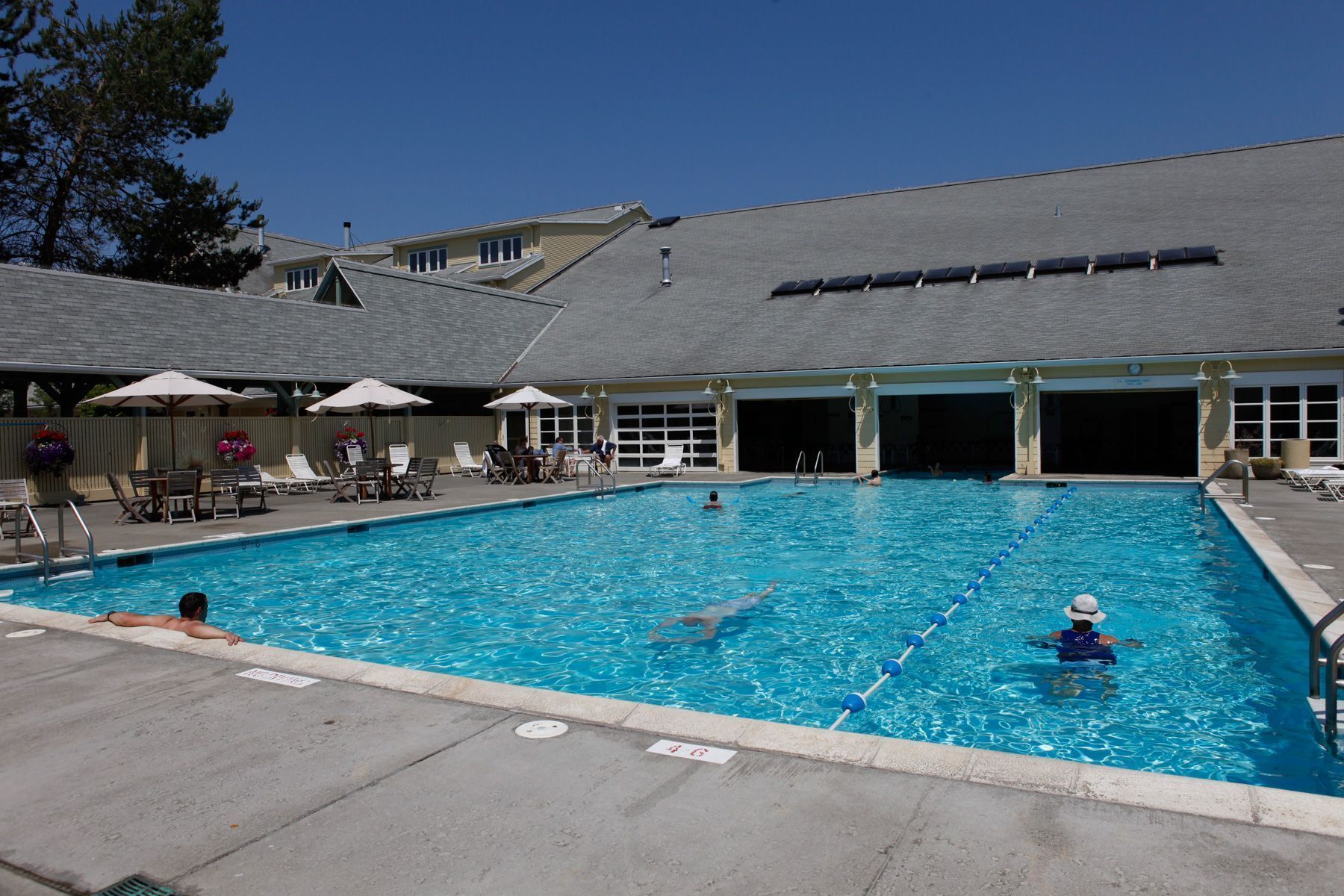 A swimming pool with lounge chairs and umbrellas, a covered area with a roof, and a building with a garage entrance.