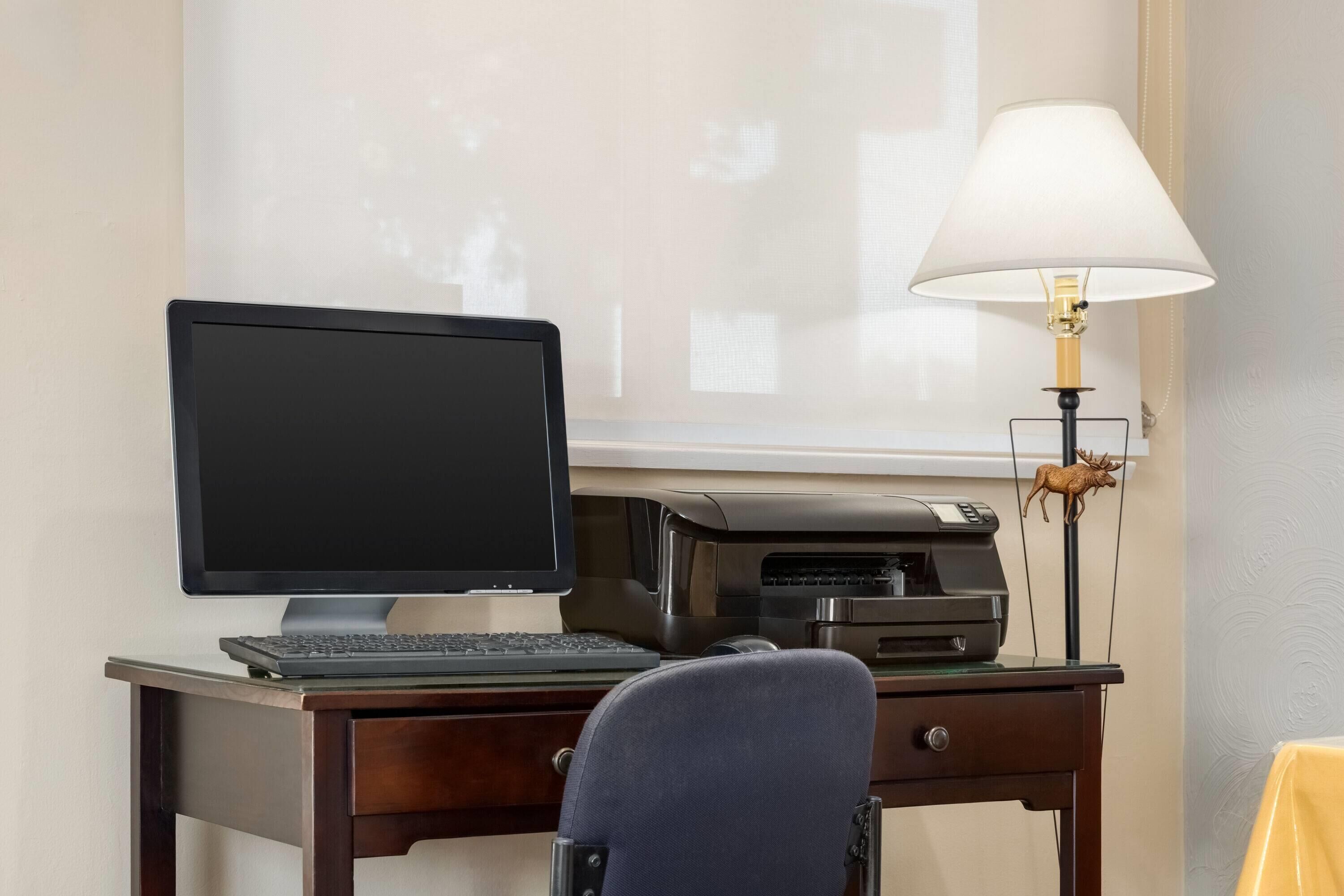 Ein Schreibtisch mit Computerbildschirm, Tastatur und Drucker, eine Lampe und ein Stuhl.