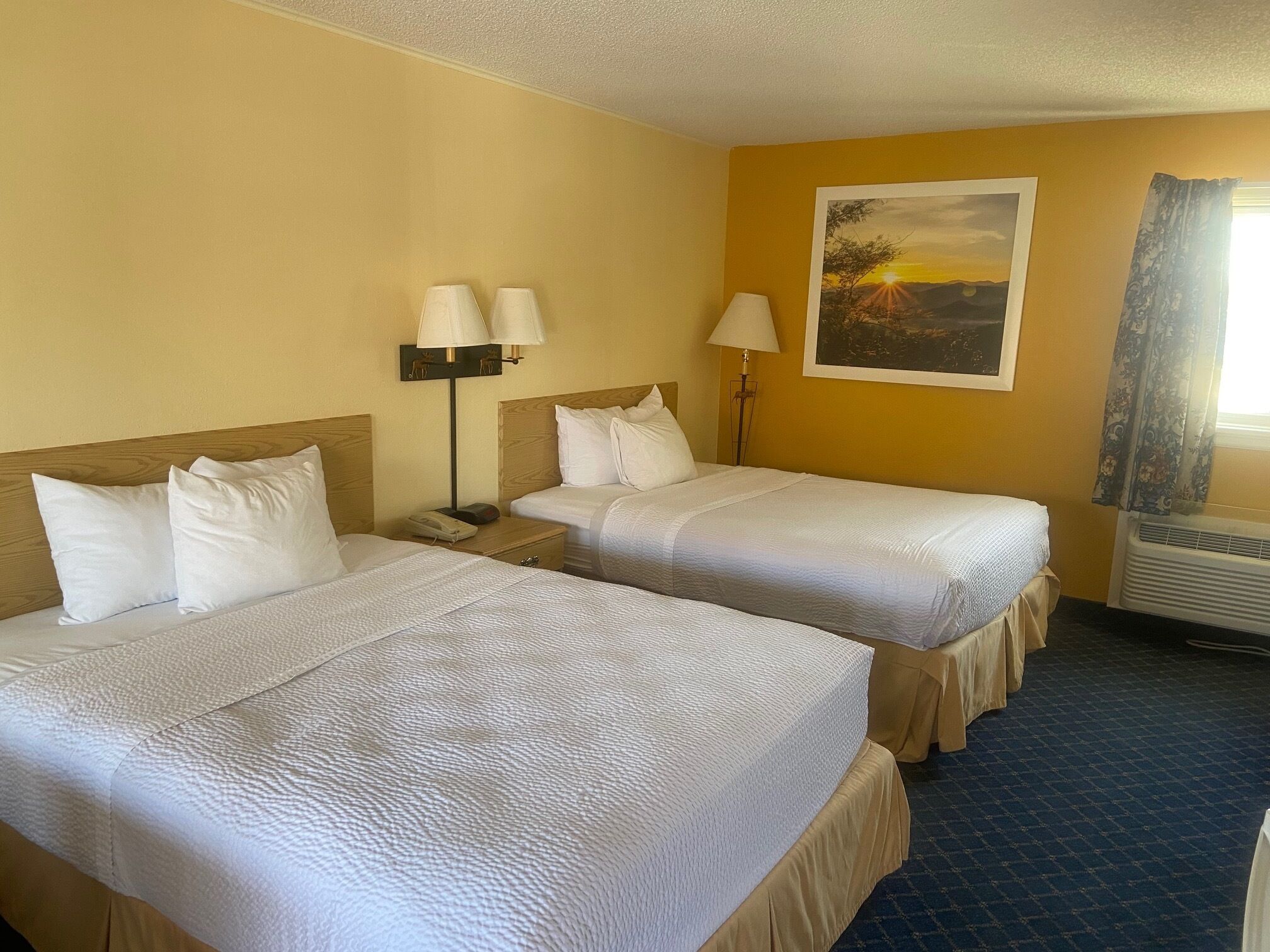 A hotel room with two beds, a nightstand with a lamp, a painting on the wall, and a window with curtains.