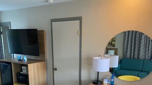 Quarto de hotel com escrivaninha de madeira, cadeira amarela, televisão e sofá verde.