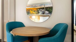 Quarto com espelho redondo refletindo uma paisagem, duas cadeiras verde-água e uma mesa de madeira.