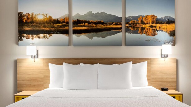 Quarto de hotel com uma cama grande, roupa de cama branca, cabeceira de madeira e três quadros de paisagens emolduradas sobre a cama.