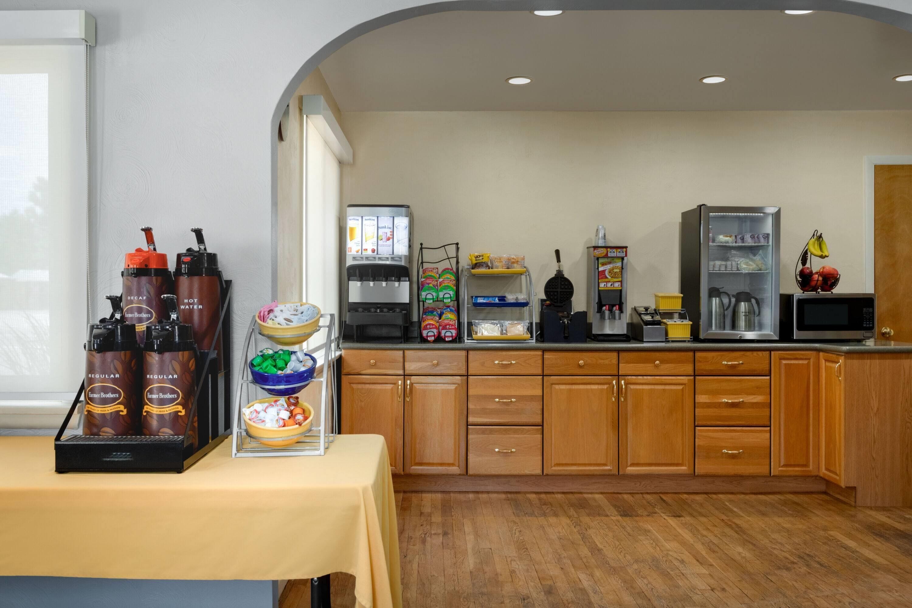 Una cocina con estación de café, microondas, refrigerador y diversos alimentos.