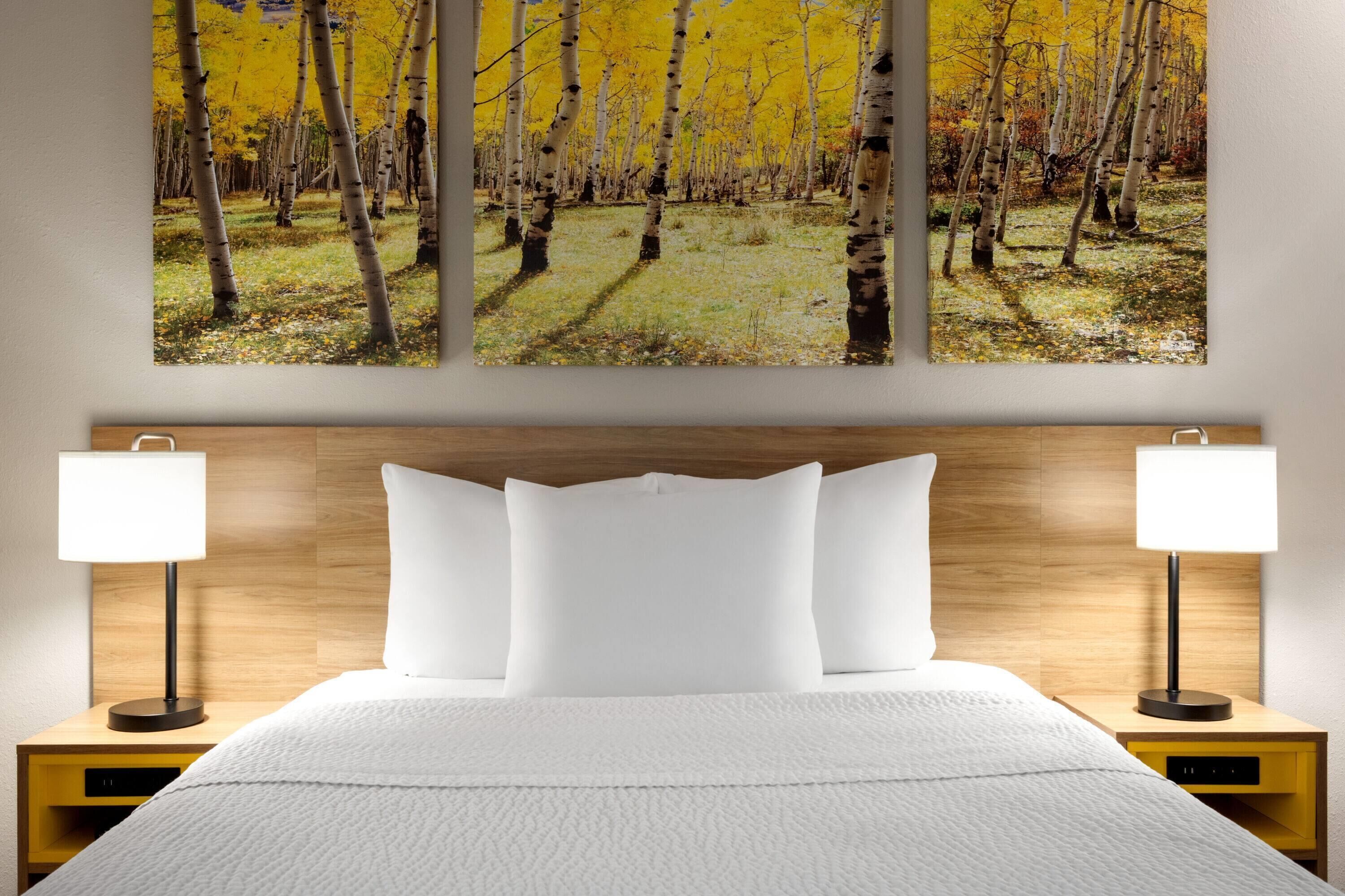 A bedroom with a large bed, two bedside tables with lamps, and a wall-mounted painting of a forest.