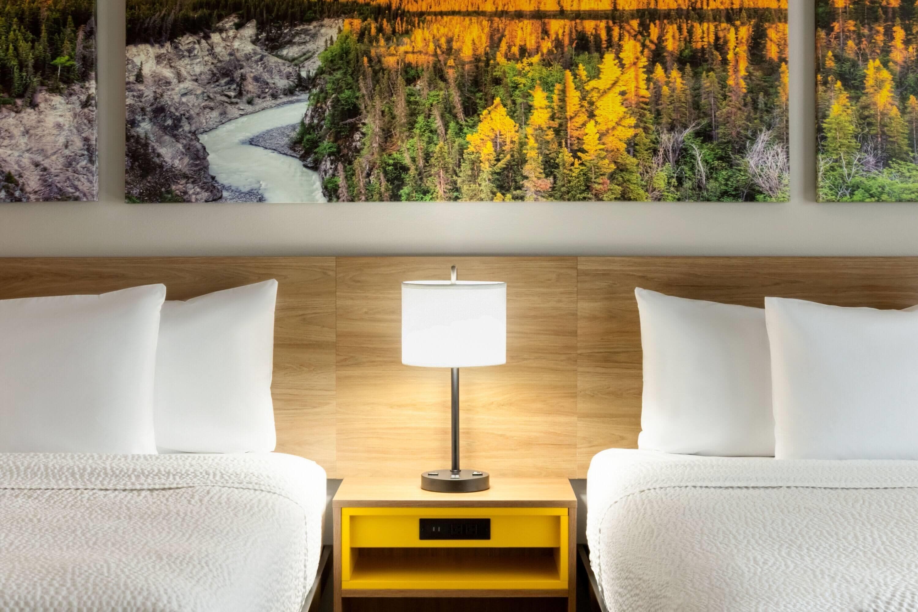 Une chambre d’hôtel avec deux lits, une table de chevet avec une lampe et un tableau représentant une forêt et une rivière, accroché au mur.