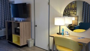 Quarto de hotel com televisão, mesa com luminária, cadeira e um quadro na parede.