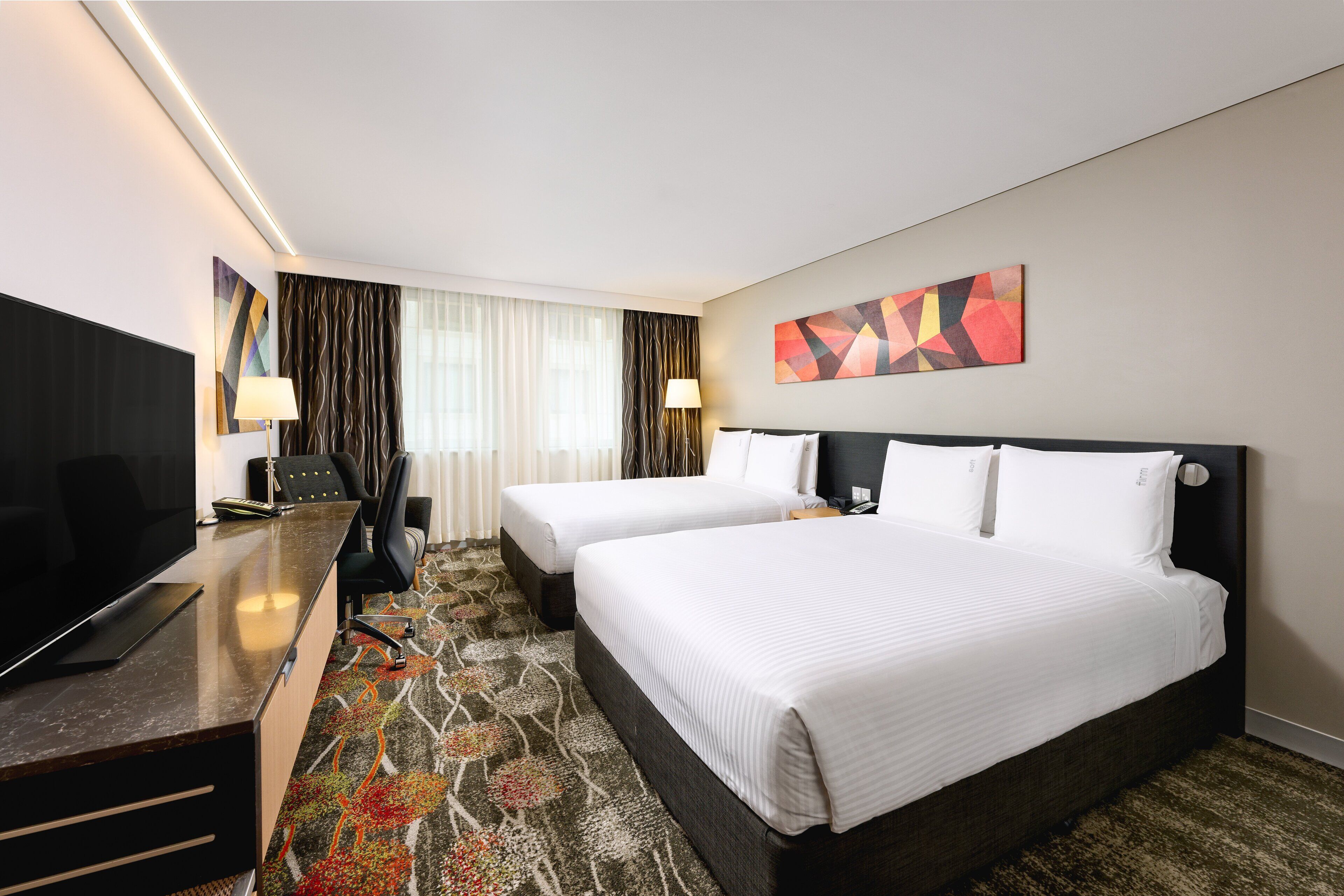 Quarto de hotel com duas camas, uma mesa com televisão, uma cadeira e piso carpeteado.