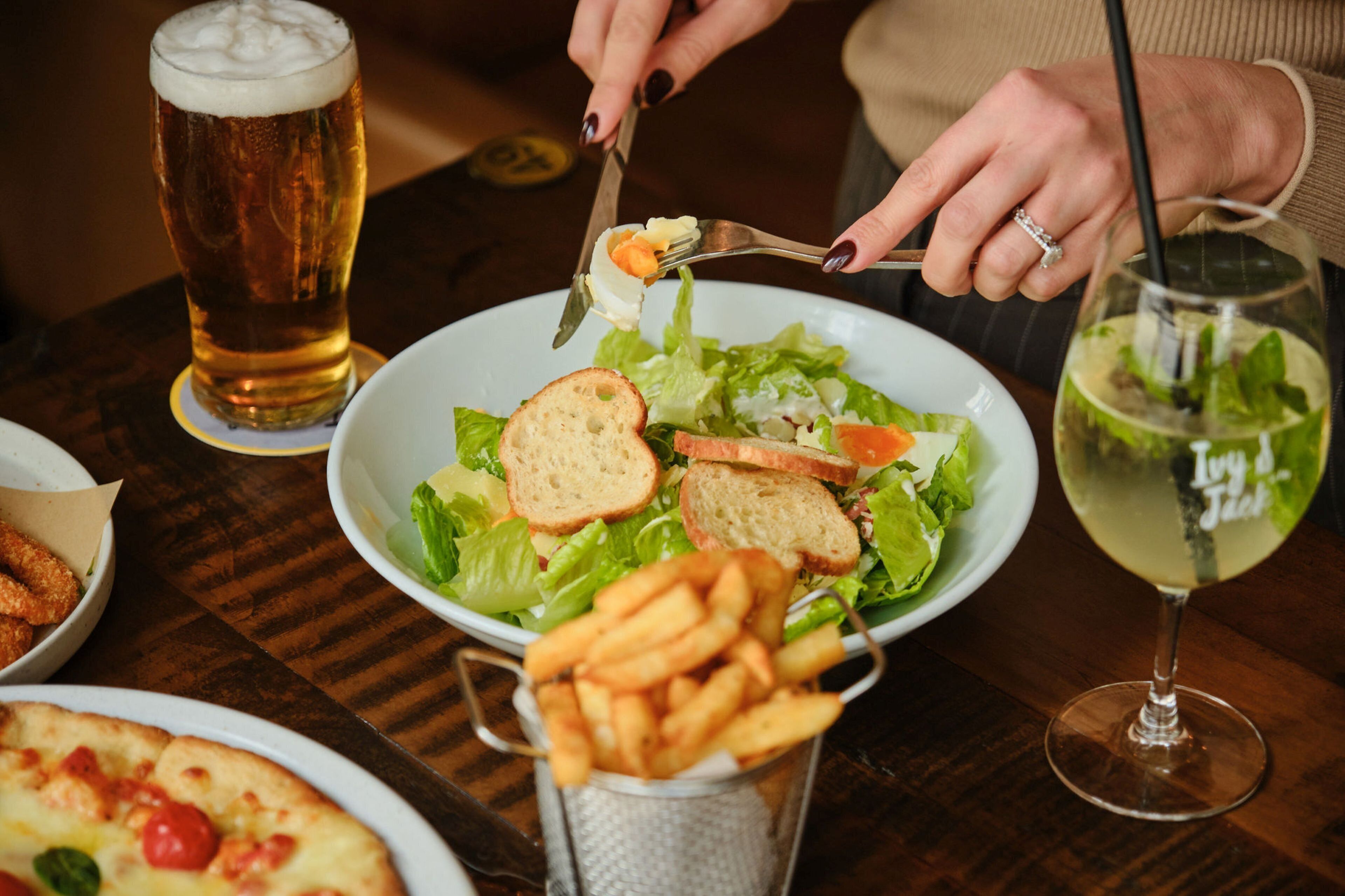 Una mesa de comedor con un plato de ensalada, una cerveza, una copa de vino blanco y una cesta de papas fritas.