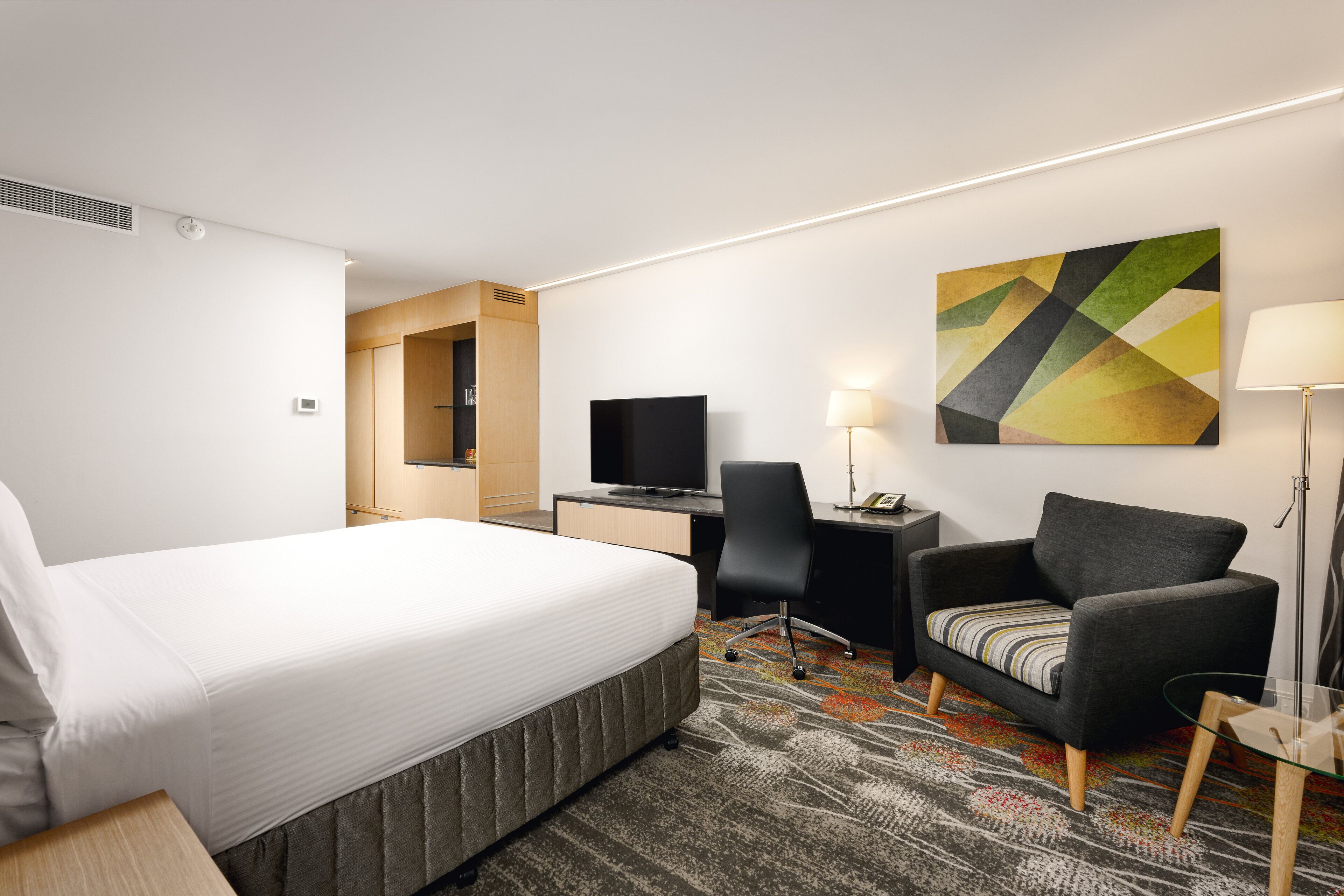 A hotel room with a bed, desk, chair, TV, lamp, and artwork.