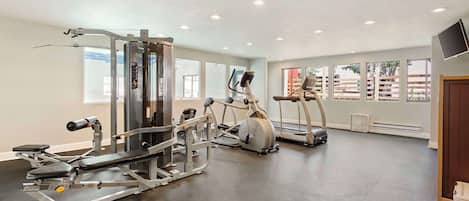 Een sportschool met diverse fitnessapparaten, grote ramen en een flatscreen aan de muur.