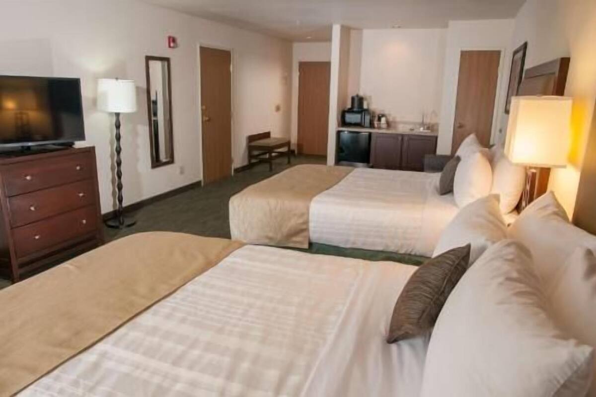 A hotel room with two beds, a desk, a chair, a television, and a kitchenette.