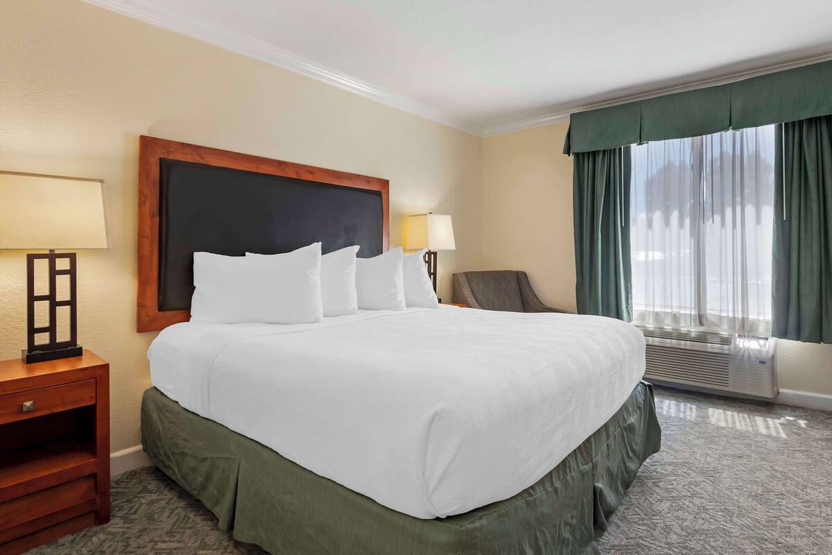 A hotel room with a large bed, two bedside lamps, a chair, a window with curtains, and a wall-mounted air conditioner.