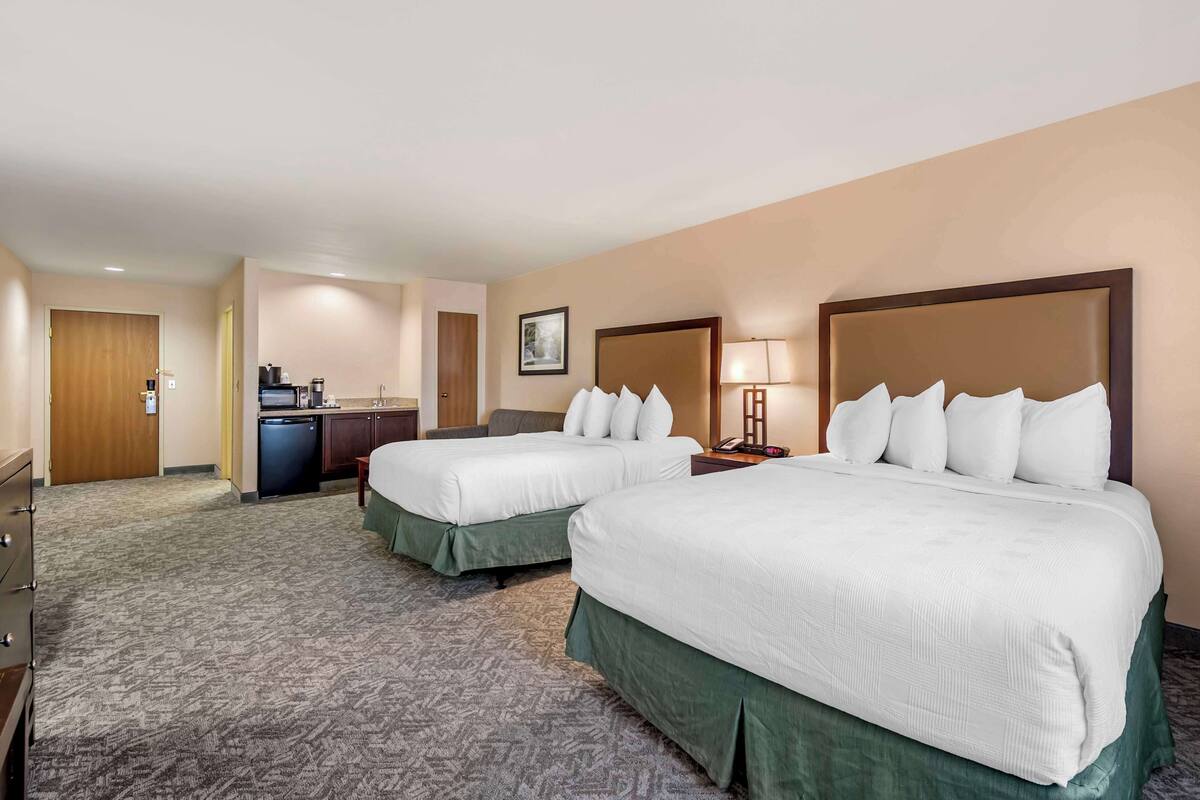 A hotel room with two beds, a desk, a chair, a lamp, and a small kitchenette.