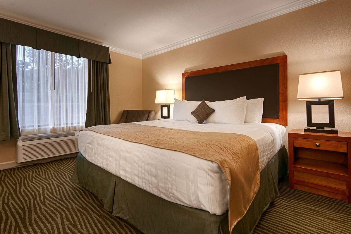 A hotel room with a large bed, two bedside lamps, a nightstand, a window with curtains, and a wall-mounted air conditioning unit.
