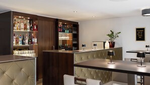 Zona de bar amb un armari de fusta ple de botelles i gots, una barra de pell amb textura i una zona de seients amb cadires i taules amb coixins.