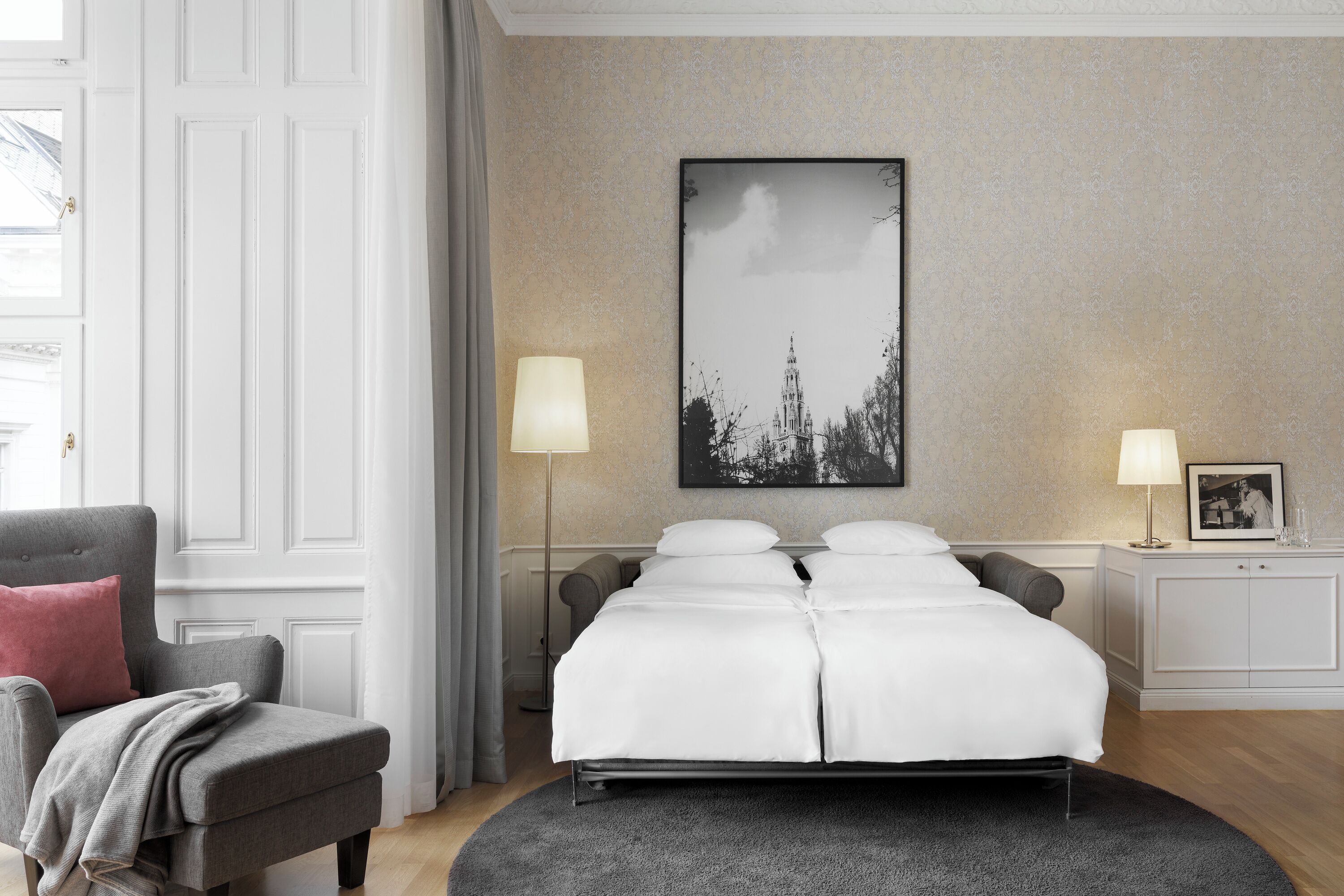 Hotelkamer met een tweepersoonsbed, twee bedlampjes, een stoel met een plaid, een ingelijst schilderij op het nachtkastje en een groot kunstwerk aan de muur.