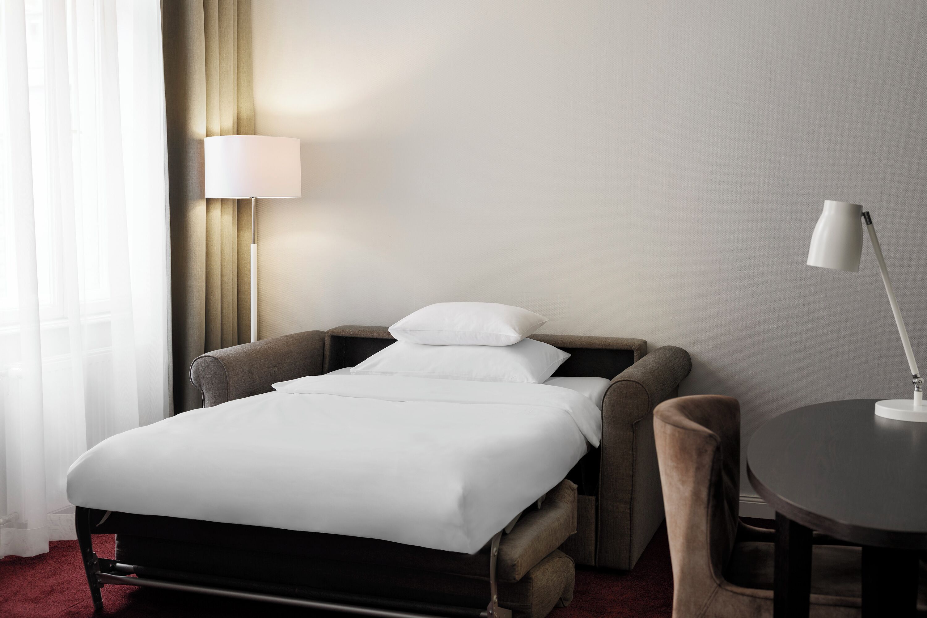A neatly made bed with white linens, a bedside lamp, and a chair next to a small table.