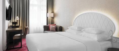 A hotel room with a large bed, a desk, a chair, a TV, and a lamp.