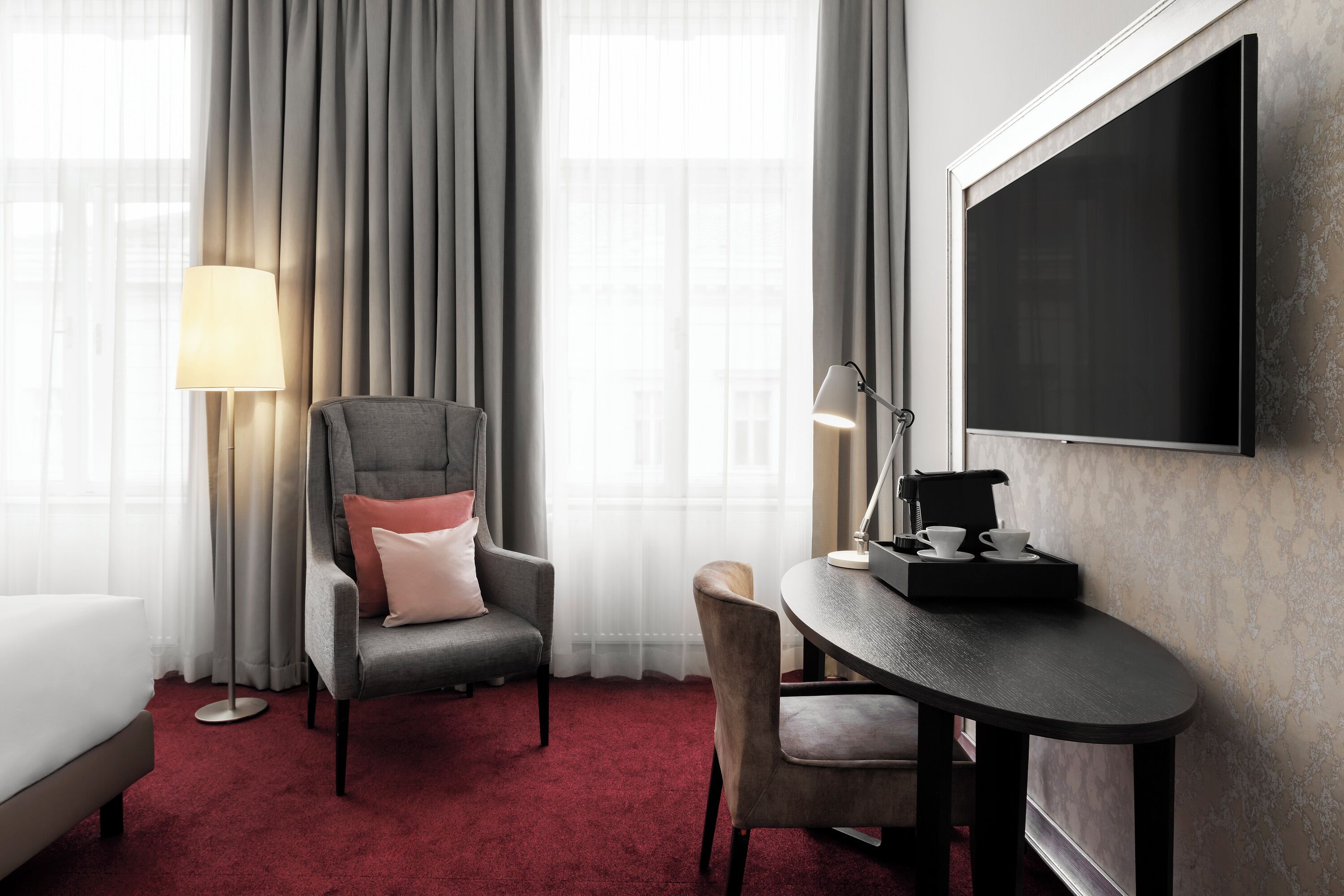 Habitación de hotel con un sillón gris, una mesita redonda con una bandeja, un sofá blanco y un televisor de pantalla plana grande montado en la pared.
