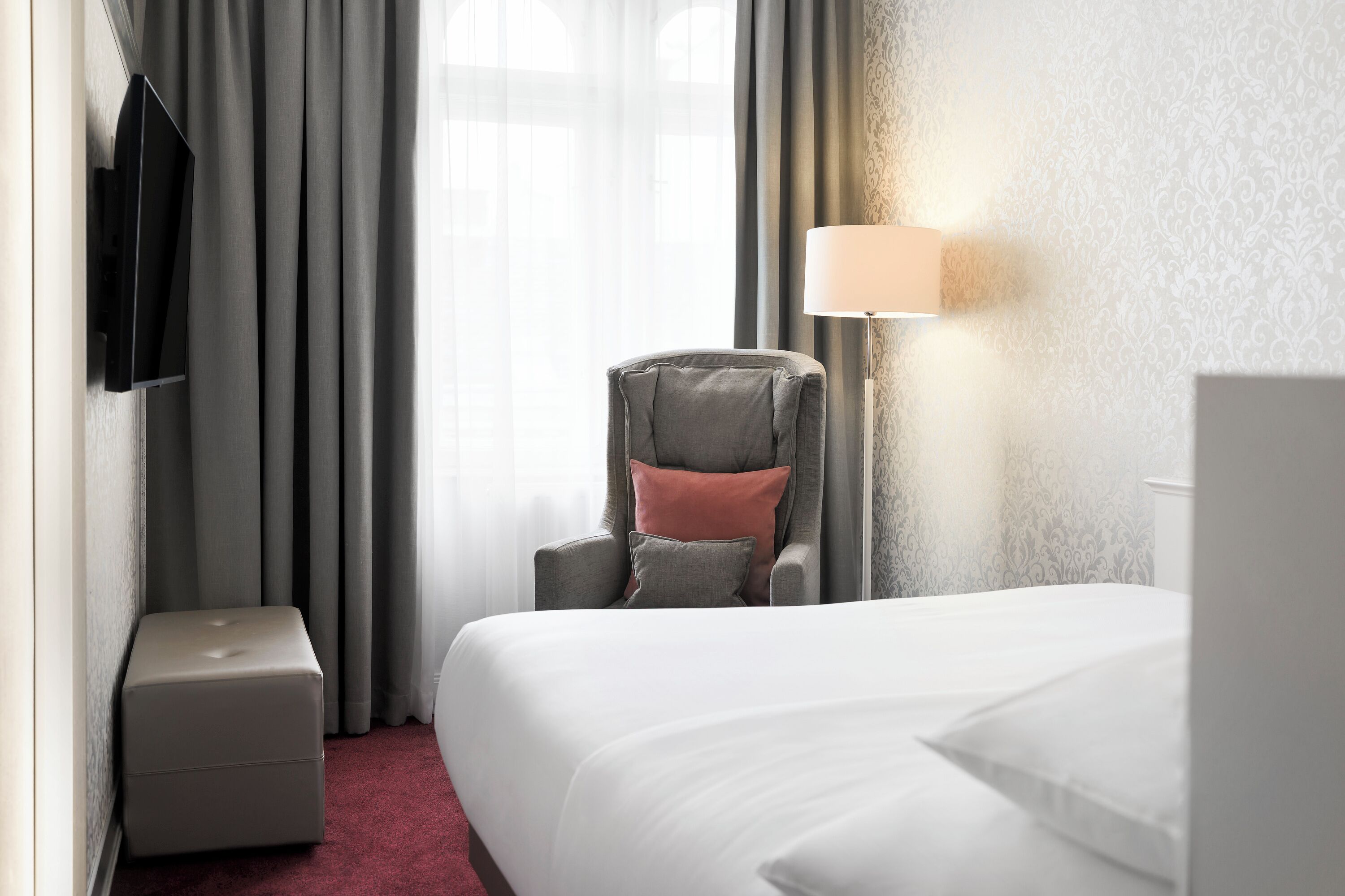 A hotel room with a bed, a chair, a small table, a TV mounted on the wall, and a lamp.