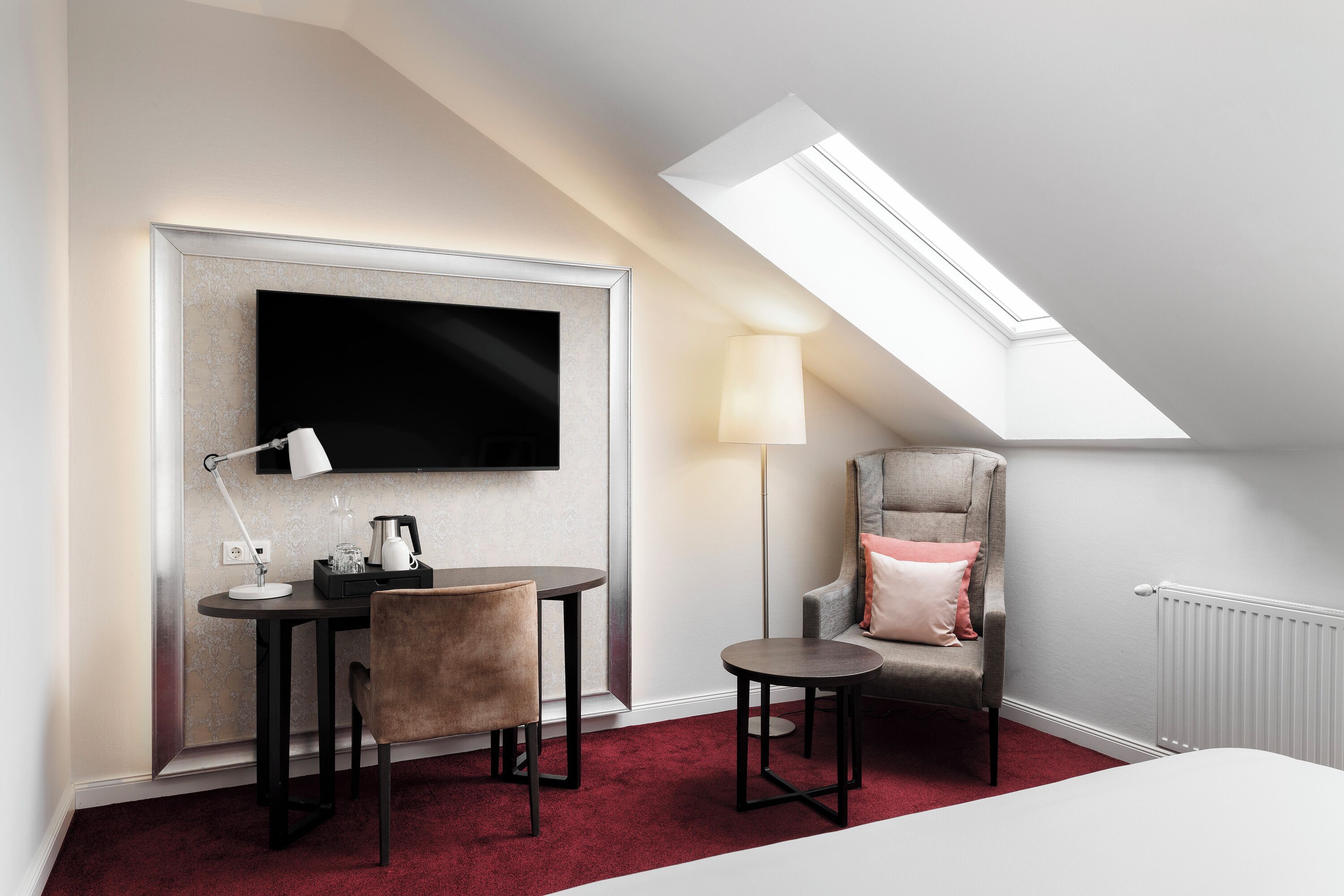A hotel room with a TV mounted on a wall, a desk with a chair, a small table, a lamp, a chair with a cushion, and a radiator.