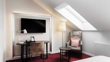A hotel room with a TV mounted on a wall, a desk with a chair, a small table, a lamp, a chair with a cushion, and a radiator.
