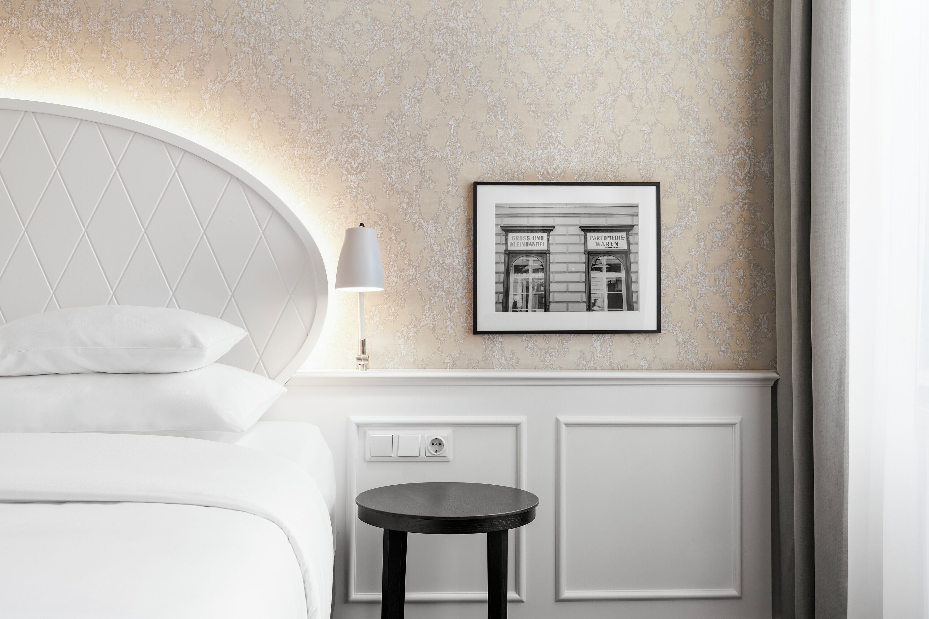 Ein ordentlich eingerichtetes Hotelzimmer mit einem Bett, einem kleinen runden Tisch, einer Wandleuchte und einem gerahmten Bild.