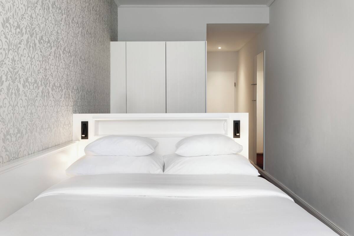 Een net opgemaakt bed met witte lakens en een hoofdbord, behang met een patroon aan de linker muur, en een witte inloopkast op de achtergrond.