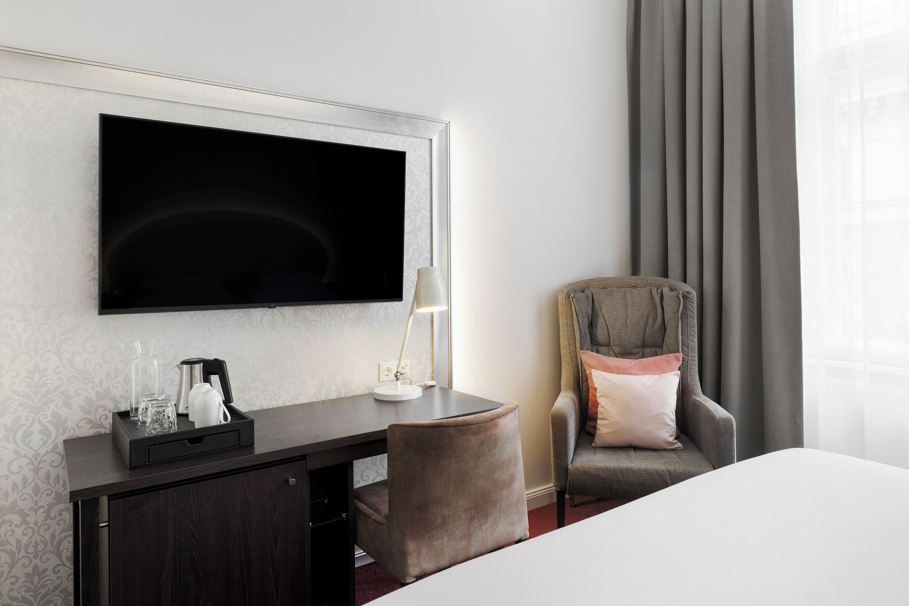 A hotel room with a flat-screen TV mounted on the wall, a desk with a lamp, a chair, and a bed.
