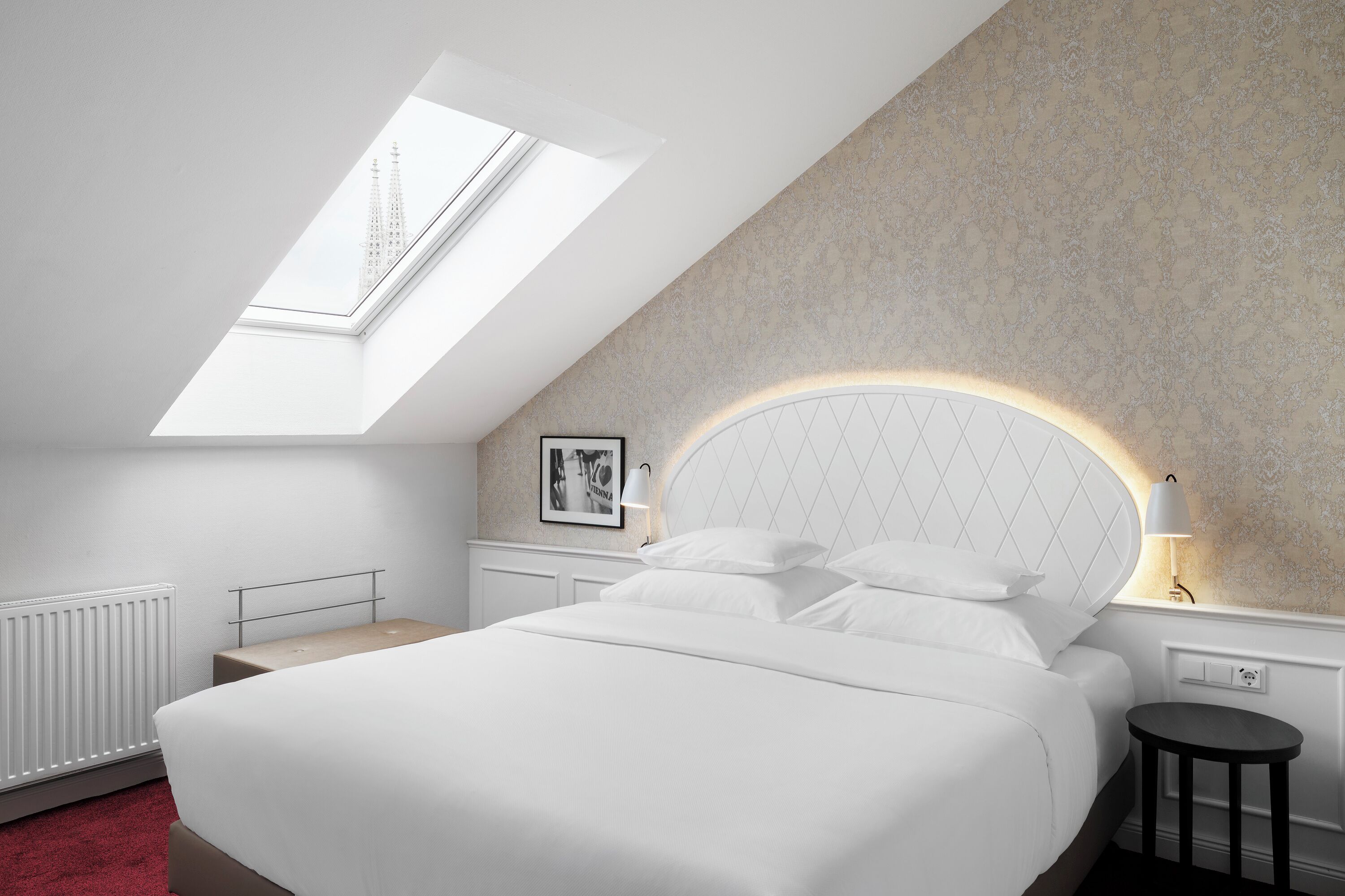 Une chambre dotée d’une lucarne, d’un lit avec une literie blanche, d’une tête de lit avec éclairage intégré, d’une table de chevet et d’un radiateur.