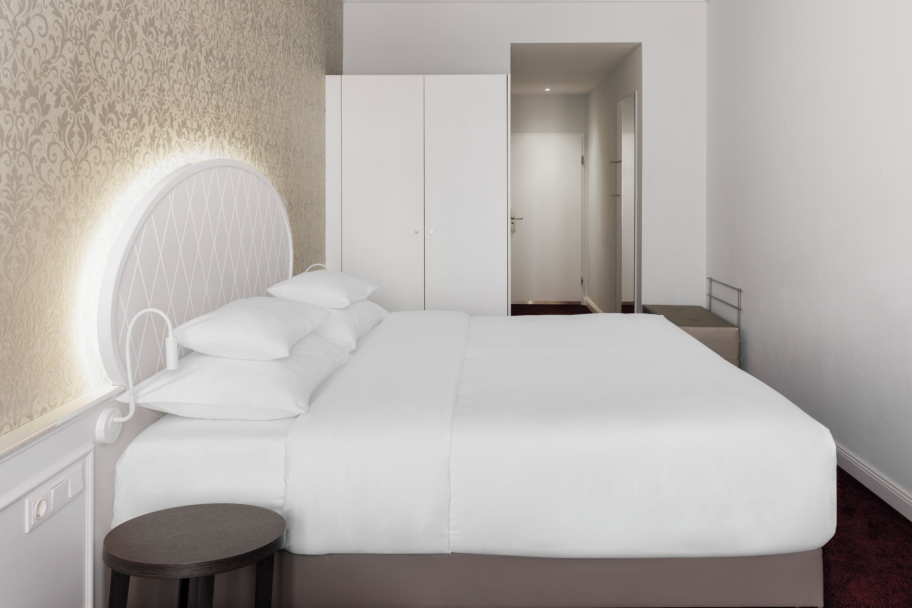 Uma cama bem arrumada com roupa de cama branca, uma mesa de cabeceira redonda e um espelho grande sobre a cama.