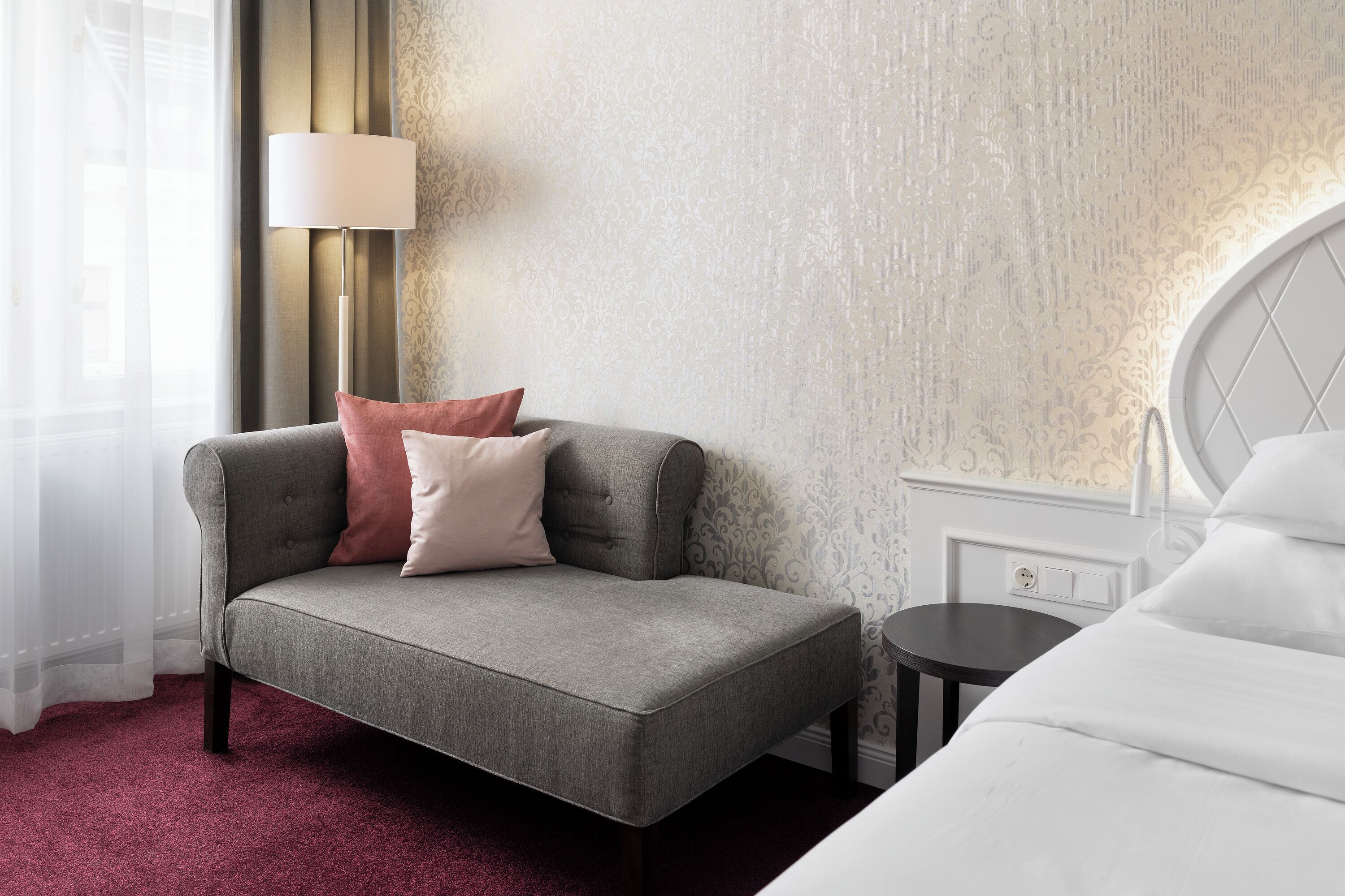 A hotel room with a grey sofa, a small round table, a white bed, and a lamp.