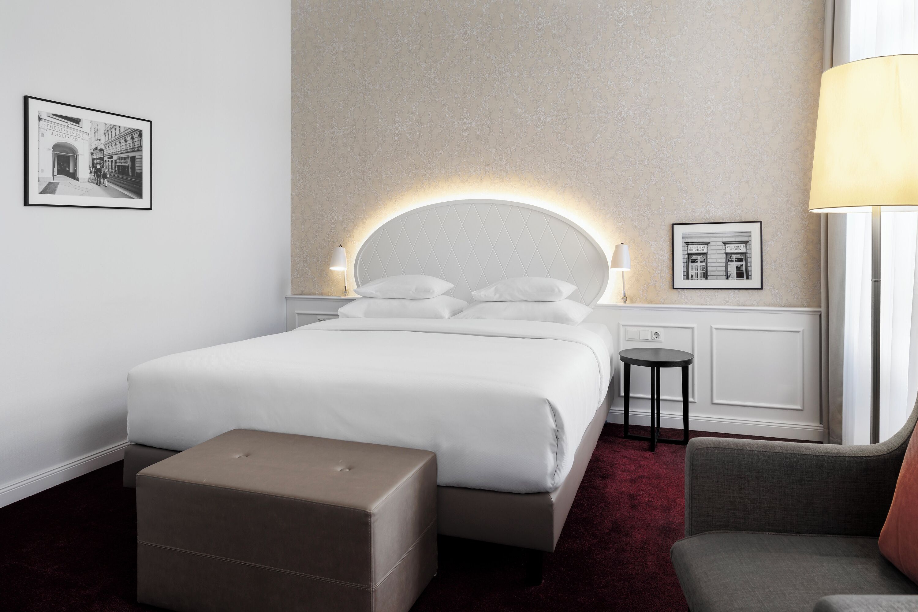 A modern hotel room with a large bed, a bedside table, a small round table, a chair, and a large window.