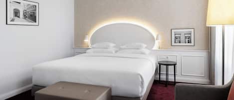 A modern hotel room with a large bed, a bedside table, a small round table, a chair, and a large window.
