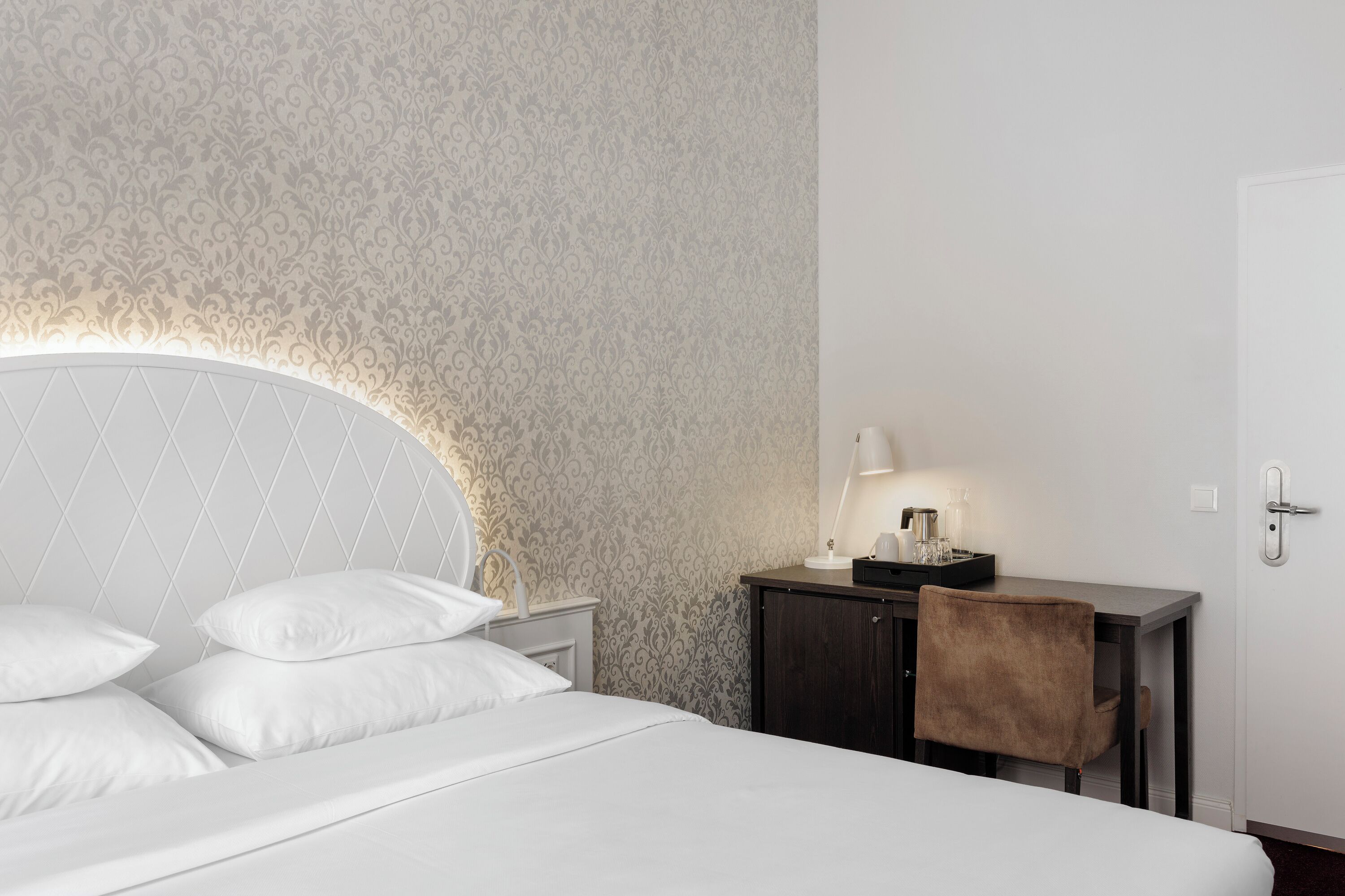 A neatly arranged hotel room with a bed, a bedside table with a lamp, a small cabinet, and a chair.