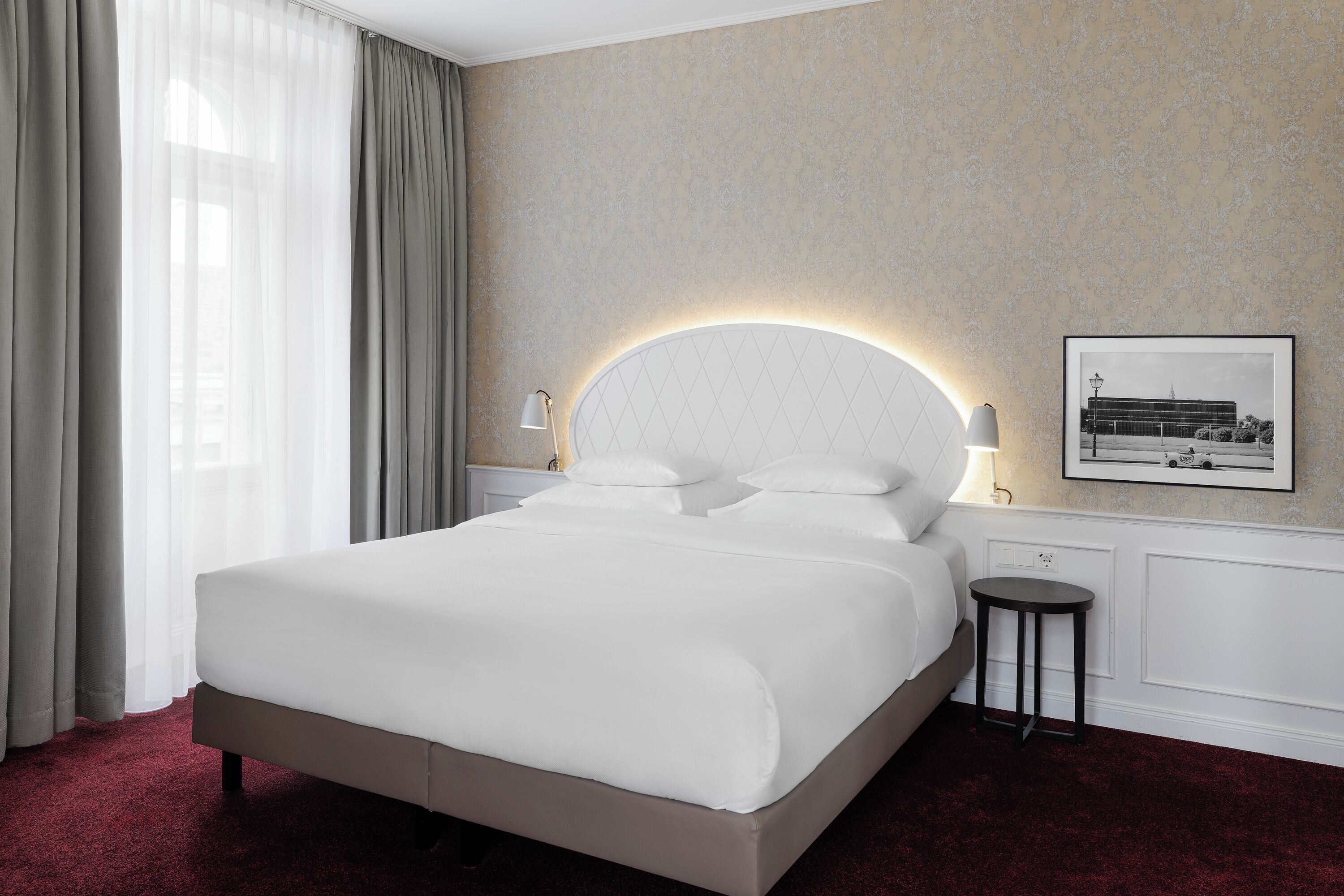 A hotel room with a large bed, a round wall-mounted light, a framed picture on the wall, and a small table beside the bed.