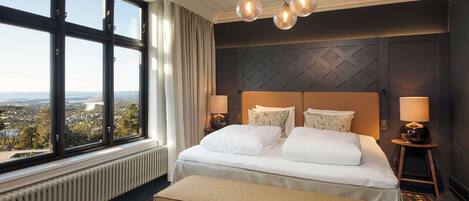 Una habitación de hotel con un ventanal grande, una cama con sábanas blancas, un cabecero con motivos geométricos y una araña.