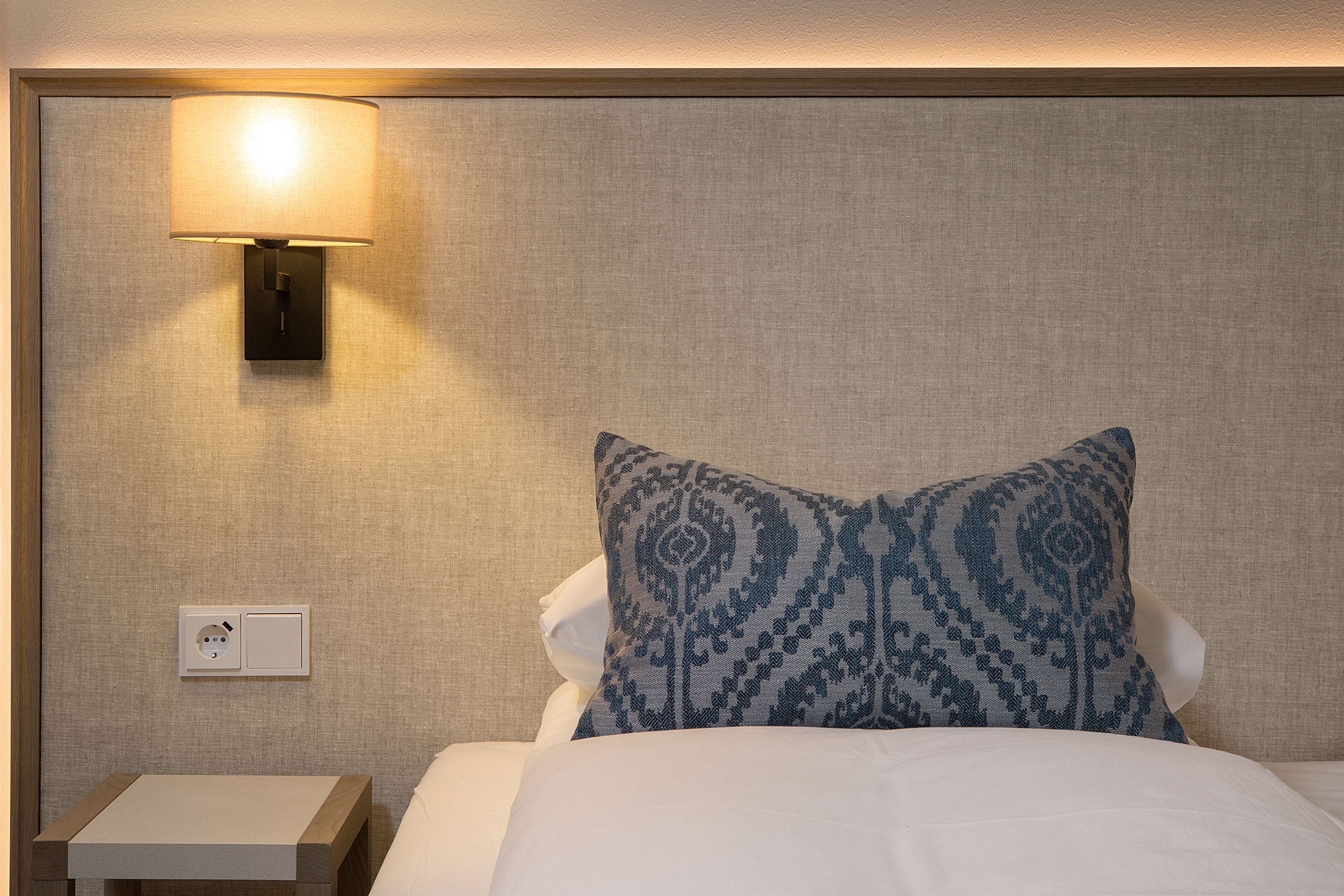 A hotel room with a bed, a nightstand, a wall-mounted lamp, and a power outlet.