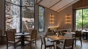 A modern dining area with round tables and chairs, large windows with a waterfall view, and a cozy seating area with sofas and a coffee table. - Scandic Holmenkollen Park (Oslo)