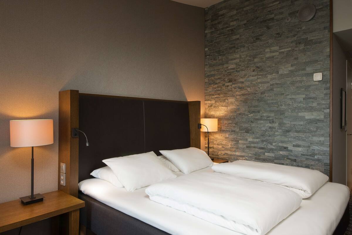 Hotellihuone, jossa on sänky, yöpöytä, lamppu ja seinä, jonka pinta muistuttaa kiveä.