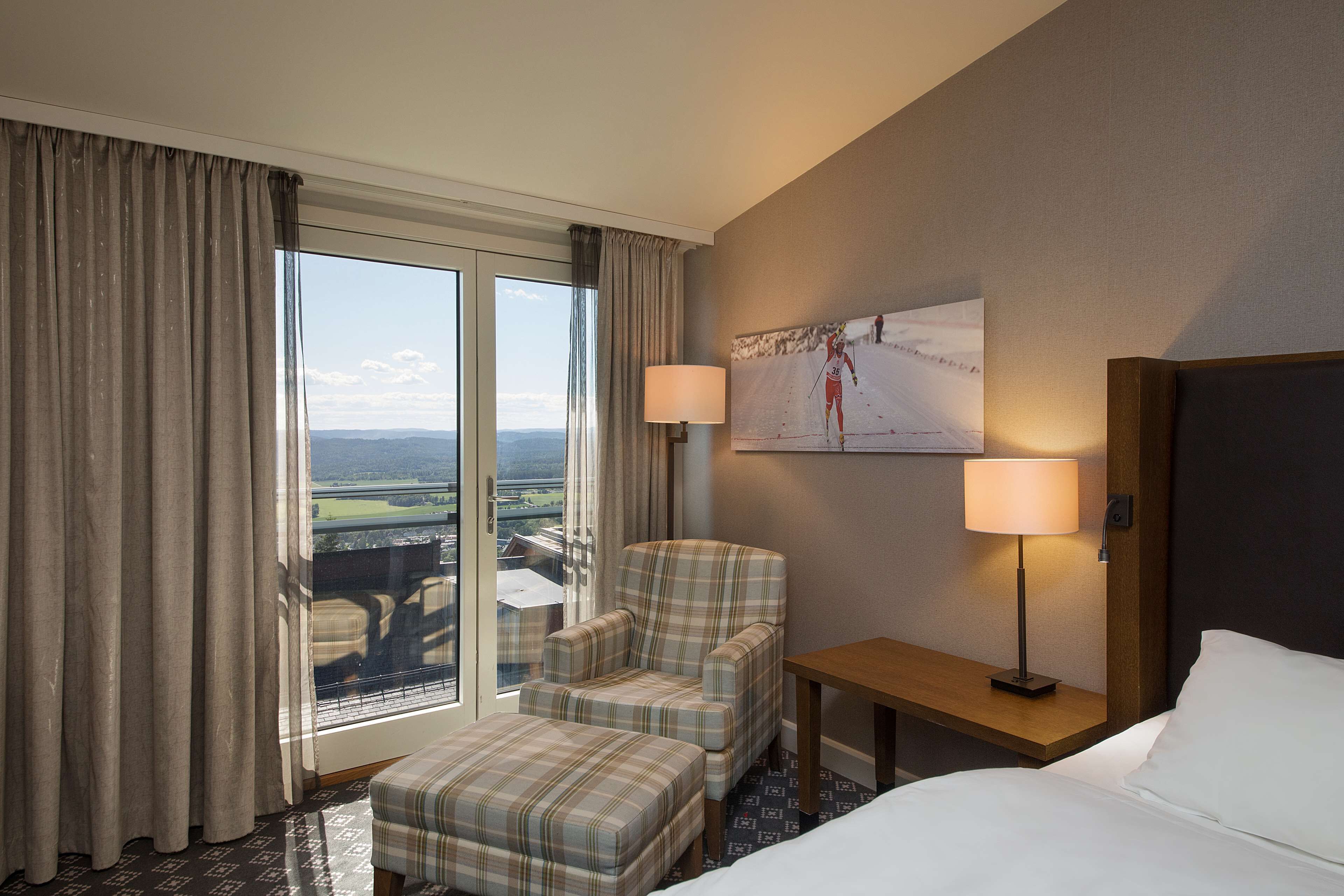 A hotel room with a bed, a chair, a small table, and a lamp. There is a large window with curtains, and a painting on the wall.