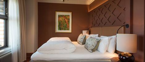 A hotel room with a large bed, a headboard with geometric patterns, a chandelier, a painting on the wall, and a window with curtains.