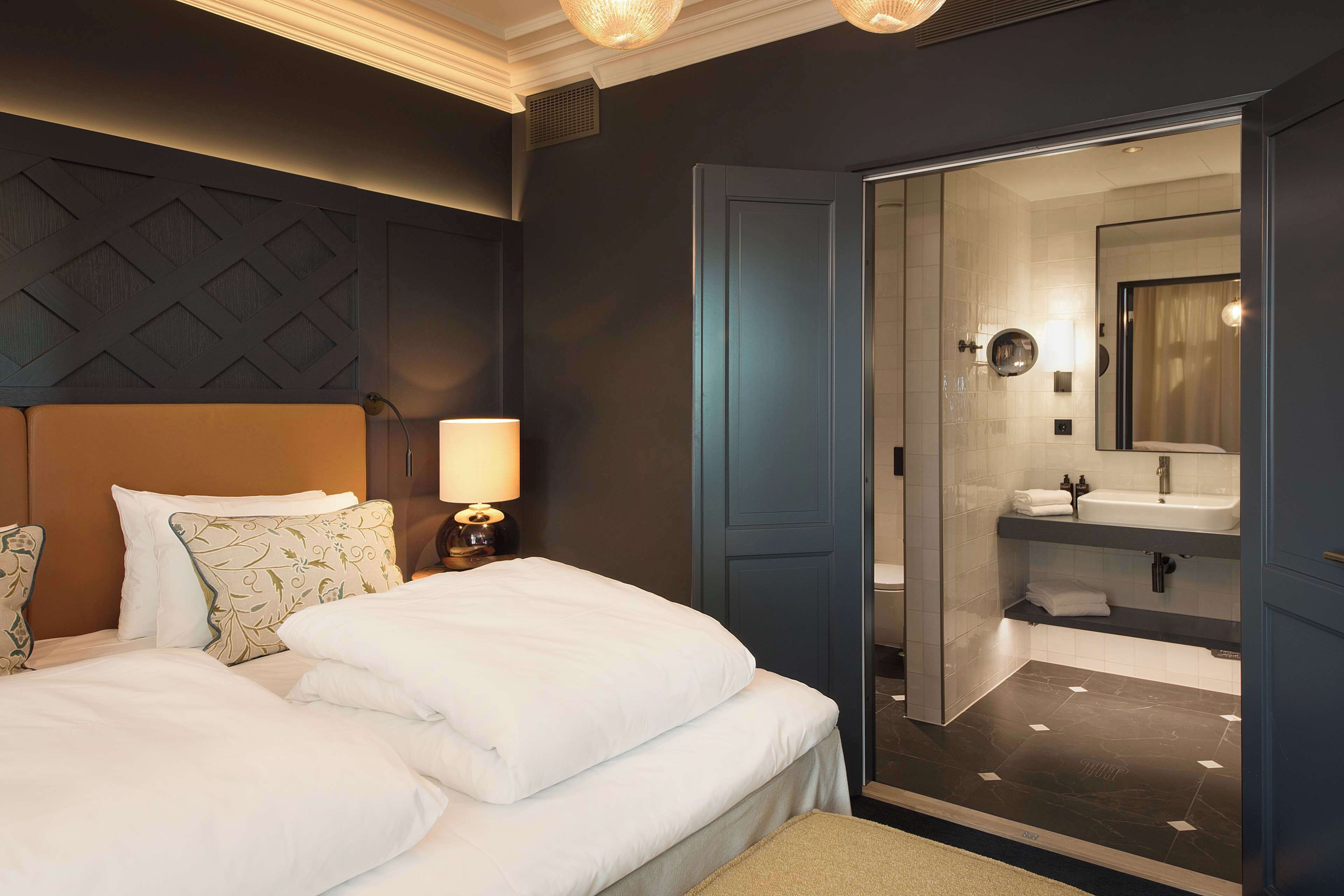 Una habitación de hotel con una cama, una mesita de noche con una lámpara, un baño con lavamanos y espejo, y una puerta que da al baño.